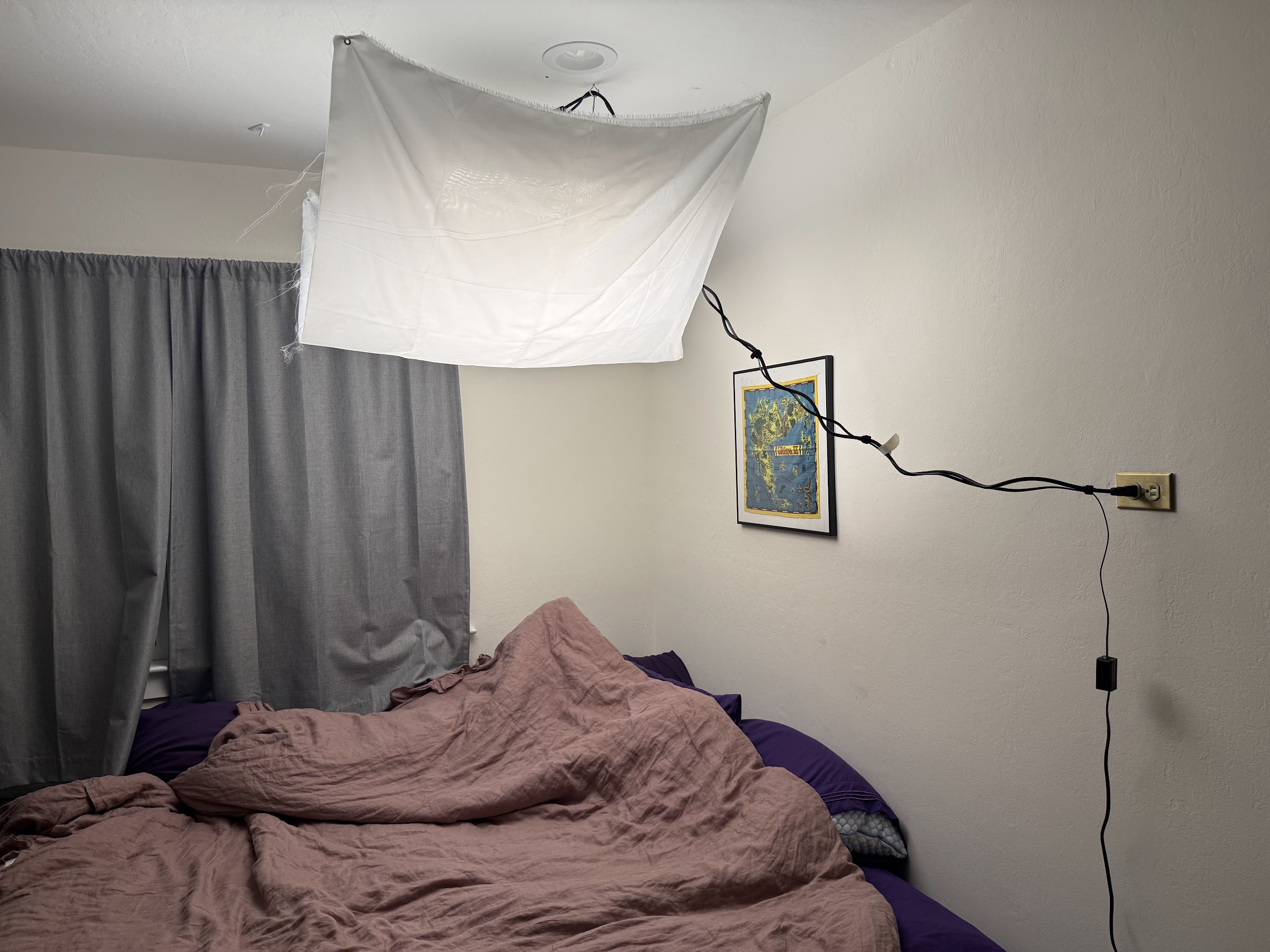 Bedroom with unmade bed, white sheet hung on ceiling, and framed map.
